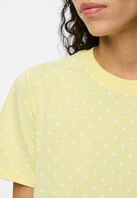 Gul t-shirt med vita prickar, kort ärm, rund halsringning och mjuk bomullstextur. Detaljer inkluderar en enfärgad kant vid halsringningen.