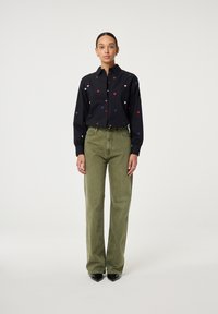 Zwarte button-up blouse met multicolor hartbrodering, gecombineerd met hoog-getailleerde olijfgroene broek en zwarte puntige hakken.