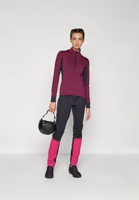 Veste zippée bordeaux avec des accents noirs, associée à un pantalon noir avec des sections roses sur le bas des jambes. Le modèle tient un casque noir.