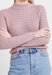 Knit sweater in light pink with a textured pattern, high neckline, and ribbed cuffs, paired with blue high-waisted jeans.