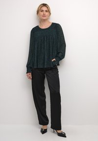 Una blusa di un verde scuro con una vestibilità comoda, dettagli arricciati sul collo e maniche lunghe. Abbinata a pantaloni di satin neri eleganti e tacchi.