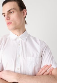 Camisa blanca de manga corta hecha de algodón ligero, con cuello button-down y un bolsillo en el pecho. Textura suave y diseño clásico.