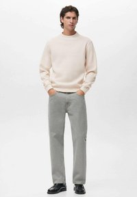 Sudadera de cuello redondo rosa claro, jeans rectos grises y zapatos negros. El modelo posa con las manos en los bolsillos contra un fondo blanco.