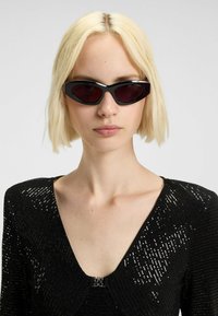 Zonnebril met een zwart en rood kleurenschema, gestroomlijnd ovale vorm en smal zwart montuur, gecombineerd met een zwarte outfit met textuur en pailletten.