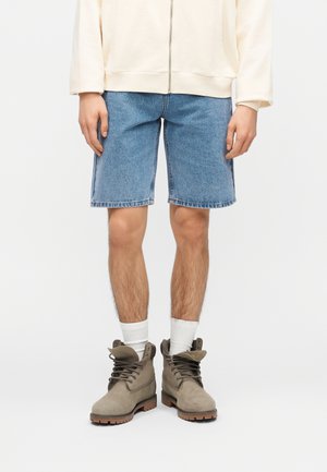 Osoba odjevena u srednje plave traper kratke hlače, bijele čarape, taupe čizme s vezicama i krem jaknu na patentni zatvarač, ruke opuštene uz bokove.