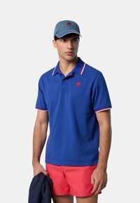 Polo blu con logo rosso, colletto a contrasto rosso e bianco. Abbinato a pantaloni corti rossi, tiene una giacca blu navy e indossa un cappellino di jeans.