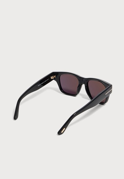 Lunettes de soleil rectangulaires noires avec verres foncés et détails dorés sur les branches, vues en angle arrière sur un fond clair.