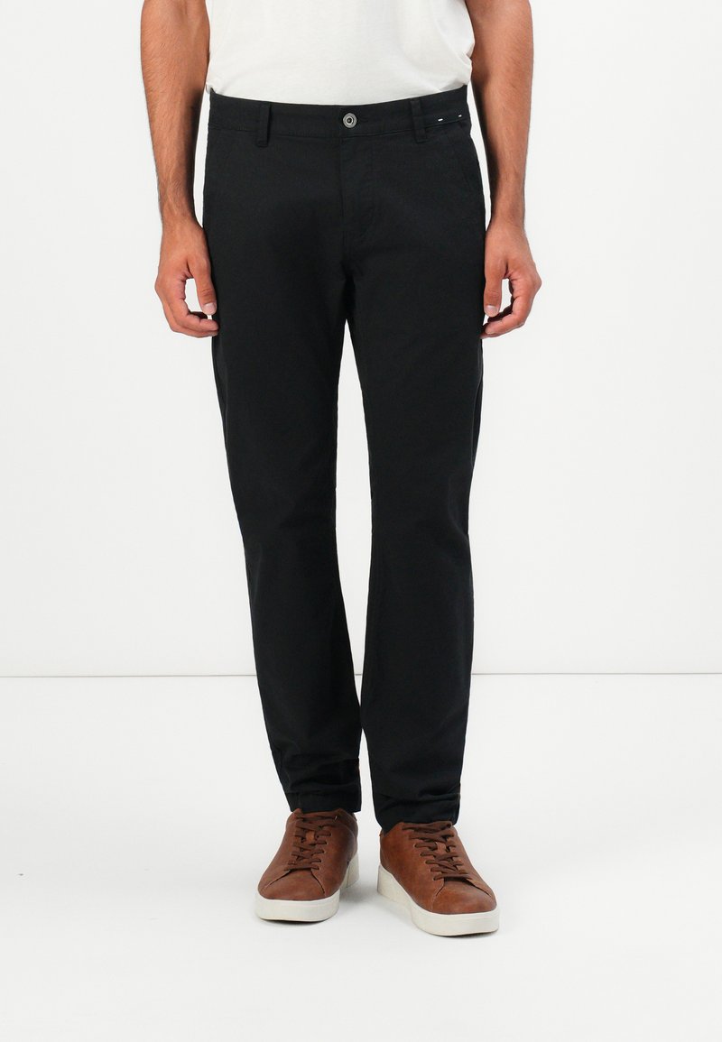 Mand iført slim-fit sorte bukser og brune sneakers står imod en ren hvid baggrund.