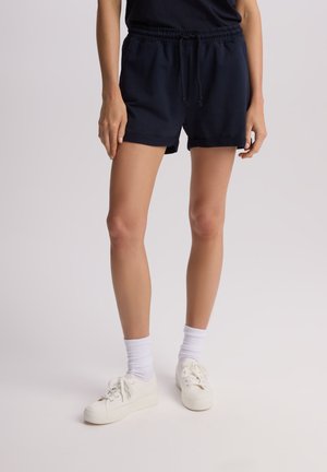 Granatowe szorty bawełniane z elastycznym paskiem i sznurkiem do regulacji. Charakteryzują się luźnym fasonem i podwiniętym dołem, zestawione z białymi trampkami i skarpetkami.
