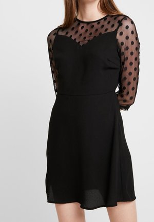 Czarna sukienka z przezroczystymi, siateczkowymi, długimi rękawami w groszki oraz prześwitującą górną częścią na klatce piersiowej, dopasowanym gorsetem i rozkloszowanym dołem, noszona przez kobietę.