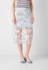 Jupe en tulle floral bleu clair avec des sequins blancs et argentés, laissant entrevoir les jambes. Associée à des sandales ouvertes bleu clair. Motif floral vertical.