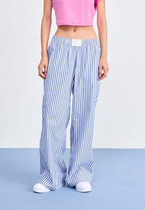 Kobieta ubrana w luźne niebiesko-białe pionowo paskowane spodnie z białą metką na pasie i białe trampki, stojąca na niebieskiej podłodze.