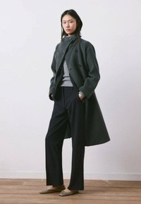 Mujer joven de pie con las manos en los bolsillos, vistiendo un abrigo gris oscuro, un cuello alto gris, pantalones negros y zapatos planos beige sobre un suelo de madera.