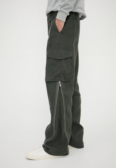 Cargo pants in dark green fabric, featuring multiple pockets, side zippers, and a relaxed fit, paired with white sneakers.