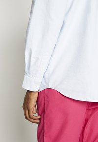 Chemise bleu clair à boutons avec manches longues et texture subtile, associée à un pantalon rose. Main visible saisissant le tissu sur le côté.