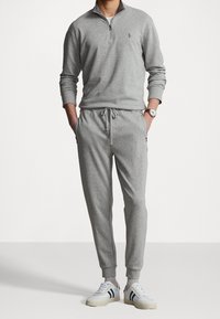 Grå zip-up tröja och matchande joggers, tillverkade av mjukt tyg, med dragsko i midjan och ribbade anklar. Vita sneakers med gröna accenter.