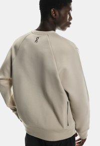 Sweatshirt bej cu o finisare texturată, având un guler ribban, mâneci raglan și un logo negru pe spate. Detaliu cu buzunar lateral cu fermoar.