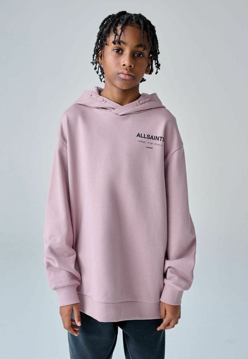 Jeune personne aux cheveux tressés portant un sweat à capuche ample de couleur violet clair avec le texte « ALLSAINTS », debout devant un fond uni.