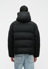 Juoda pūkinė striukė su aukštu apykakle ir gaubtu, su siuvinėtomis dalimis; derinama su šviesiai mėlynomis džinsomis, kurios rodo kišenes.