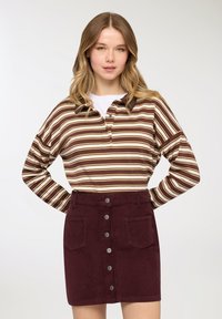 Giovane donna con capelli biondi ondulati che indossa una maglia a maniche lunghe a righe marroni e beige e una gonna bordeaux abbottonata.