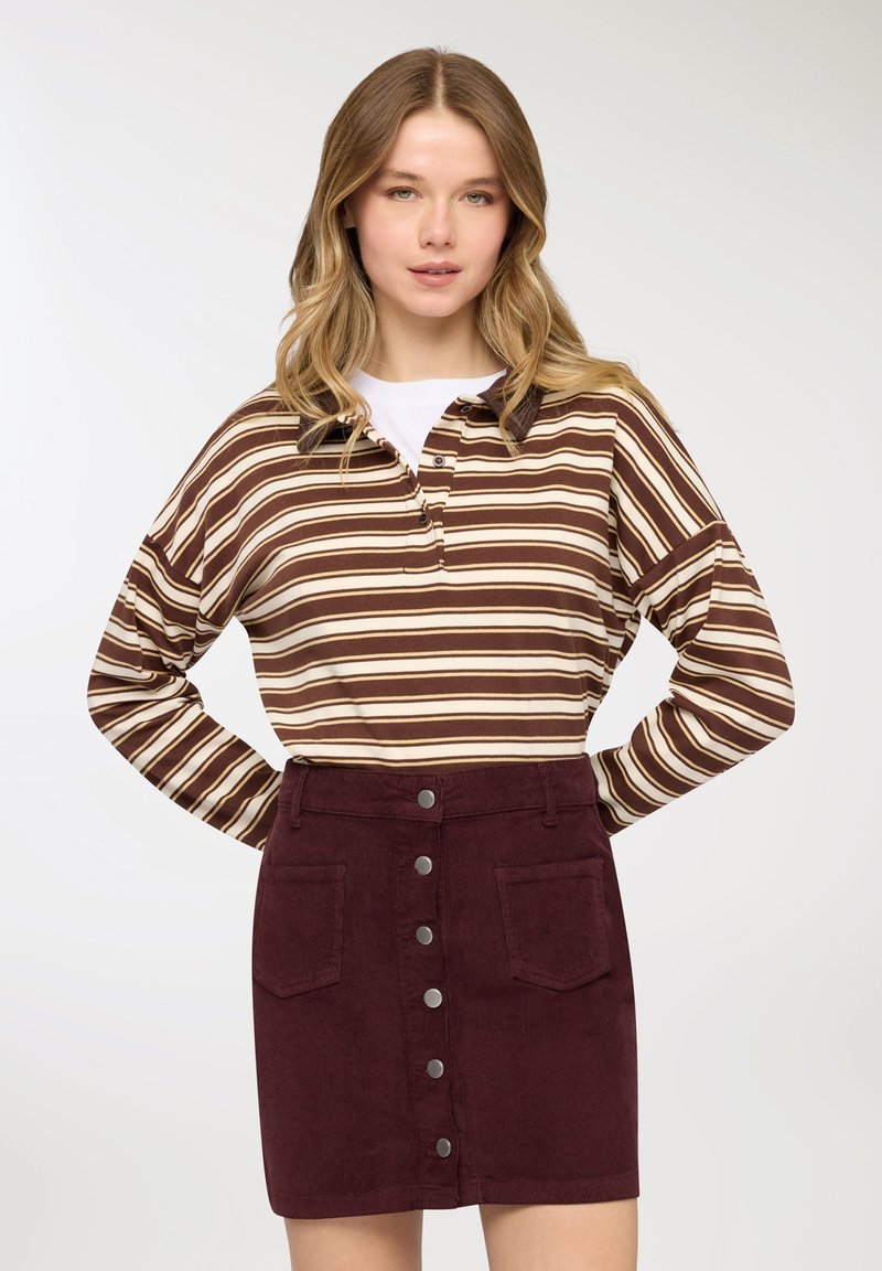 Giovane donna con capelli biondi ondulati che indossa una maglia a maniche lunghe a righe marroni e beige e una gonna bordeaux abbottonata.