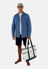 Spijkerblouse, witte t-shirt, donkere shorts, met een grote crèmekleurige tote bag met marineblauwe accenten en een voorvak. Model is blootsvoets.