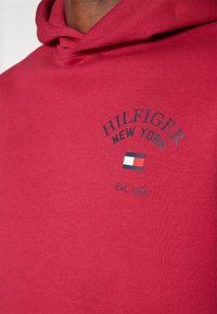 Czerwona bawełniana bluza z kapturem z przodu z logo "HILFIGER NEW YORK", małą grafiką flagi i napisem "Est. 1985" w kontrastującym granatowym kolorze.