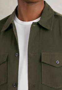 Samthemd in Dunkelgrün mit Knopfleiste, Kragen und zwei aufgesetzten Taschen. Über einem weißen Rundhals-T-Shirt getragen.