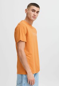Orange bomulds t-shirt med korte ærmer, rund hals, og hvid tekstlogo på fronten. Modellen har lyseblå denimshorts på.