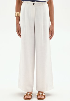 Femme portant un pantalon blanc à jambes larges, des sandales marron avec de grandes décorations en bijoux, un bracelet manchette en or et un haut rayé partiellement visible.