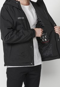 Svart vattentät jacka med huva, med ett vadderat foder och sidofickor. Inuti finns en etikett med skötselråd och symboler.