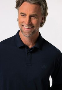 Tmavě modré polo tričko vyrobené z hladké tkaniny, s klasickým límečkem a jemně vyšitým logem na levé části hrudi.