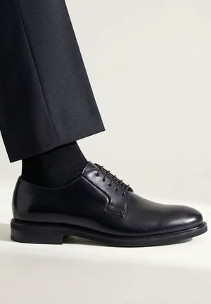 Schwarzer, glänzender Lederschuh mit Schnürsenkeln, getragen mit schwarzen Socken und dunkelgrauer formeller Hose vor hellem Hintergrund.