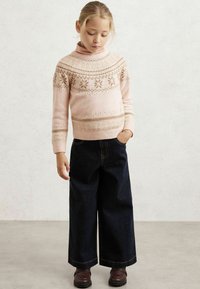 Mladé dievča s blond vlasmi, oblečené v svetlo ružovom vzorovanom svetri, tmavých širokých nohaviciach a hnedých topánkach, stojace s jednou rukou v vrecku.