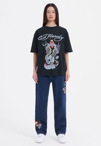 Black oversized Ed Hardy t-shirt with colorful skull and eagle graphic. Paired with blue denim pants featuring embroidered designs. White sneakers.