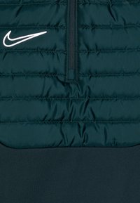 Chaqueta acolchada de color verde oscuro con un diseño de media cremallera, que presenta una textura suave y un logo de Nike en blanco en la parte superior izquierda.