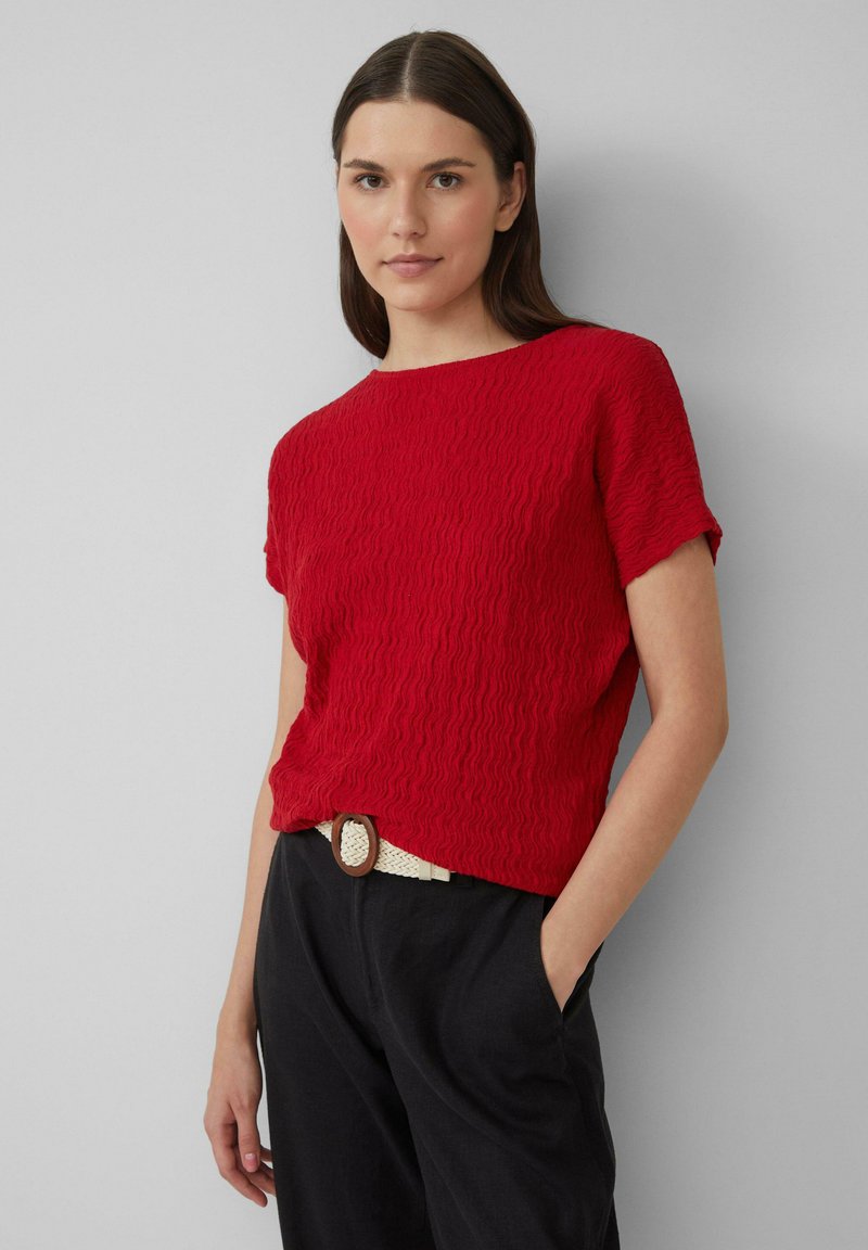 Haut rouge texturé à manches courtes avec un col rond, associé à un pantalon noir taille haute et une ceinture tressée blanche avec une boucle circulaire.
