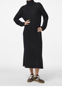 Abito nero a maniche lunghe con scollo a camicia e spacchi laterali, realizzato in materiale morbido. Abbinato a sneakers marroni e bianche.