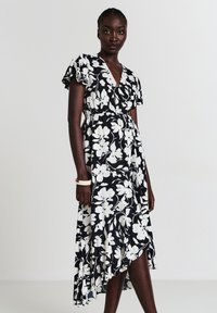 Femme portant une robe cache-cœur florale noire et blanche avec des manches volantées et un ourlet asymétrique, se tenant devant un fond clair.