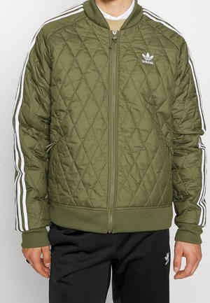 Mężczyzna noszący oliwkową pikowaną kurtkę Adidas z białymi pasami na rękawach i logo Adidas na piersi, w połączeniu z czarnymi spodniami Adidas.