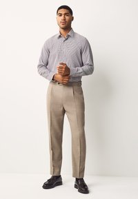 Chemise à rayures boutonnée bleu clair et blanc, associée à un pantalon beige ajusté. Le mannequin porte des chaussures noires brillantes.