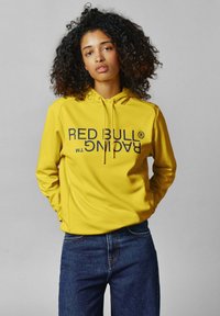Geltonas kapucinis su dideliu tamsiai mėlynu „RED BULL RACING™“ logotipu. Turi raištelį, ilgus rankovės ir minkštą audinio tekstūrą. Dėvima su mėlynomis džinsomis.