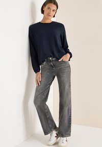 Granatowa koszulka z długim rękawem o teksturowanej tkaninie, zestawiona z wyblakłymi szarymi dżinsami o szerokich nogawkach i białymi trampkami z różowymi akcentami. Gładkie tło ściany.