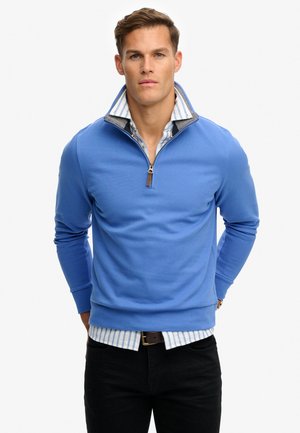 Man wearing a blue quarter-zip pullover over a striped button-up shirt, with black pants and hands behind his back.