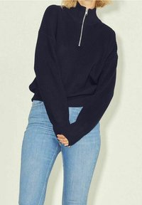 Laivastonsininen neulepullovers jossa on korkea kaulus ja hopeinen vetoketju; ominaisuuksiin kuuluu ribbikuviointi ja ylisuuria mitoituksia. Yhdistetty vaaleansinisiin farkkuihin.