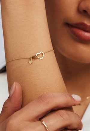 Bracelet en chaîne dorée avec un charm en forme de cœur et une petite étiquette ronde. Texture lisse, design délicat et longueur ajustable.