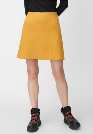 Falda de línea A amarilla en tejido texturizado, a la altura de la rodilla con costuras laterales. Combinada con botines negros con cordones y detalles en rojo.