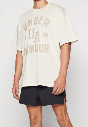 T-shirt oversized crème clair avec le texte "Under Armour" en beige, col rond, manches courtes et coupe relaxée, associé à un short noir.