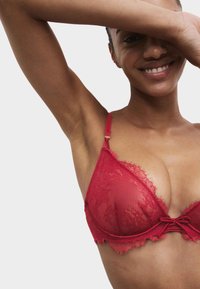 Czerwony koronkowy biustonosz o trójkątnym kształcie, z cienkimi regulowanymi ramiączkami i dekoracyjnym supełkiem na środku, zdobiony teksturowanymi kwiatowymi wzorami.