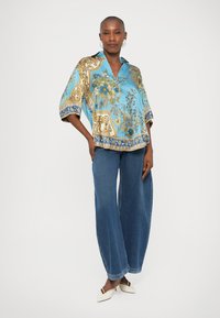 Blusa de seda em azul claro com um padrão floral em dourado, corte solto, mangas largas e colarinho. Combinada com calças de ganga de perna larga e sapatos brancos.
